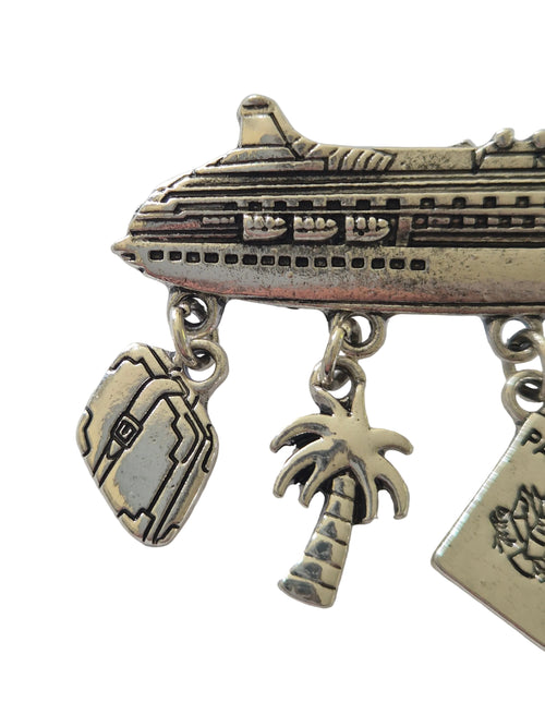 silver-colored charm brooch with airplane, palm tree, and heart design on a white background