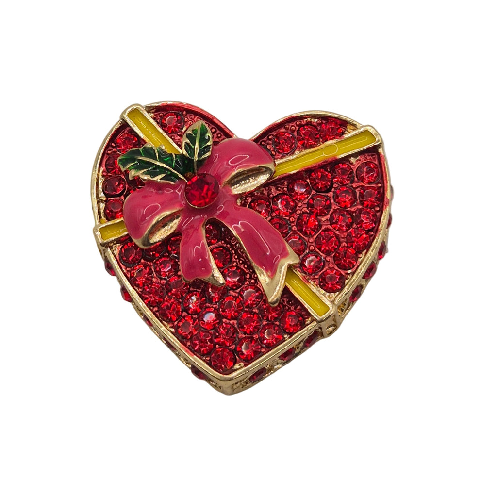 A heart-shaped brooch designed to look like a gift box, covered in red rhinestones with a pink bow, green holly leaves, and gold trim, set against a plain white background.