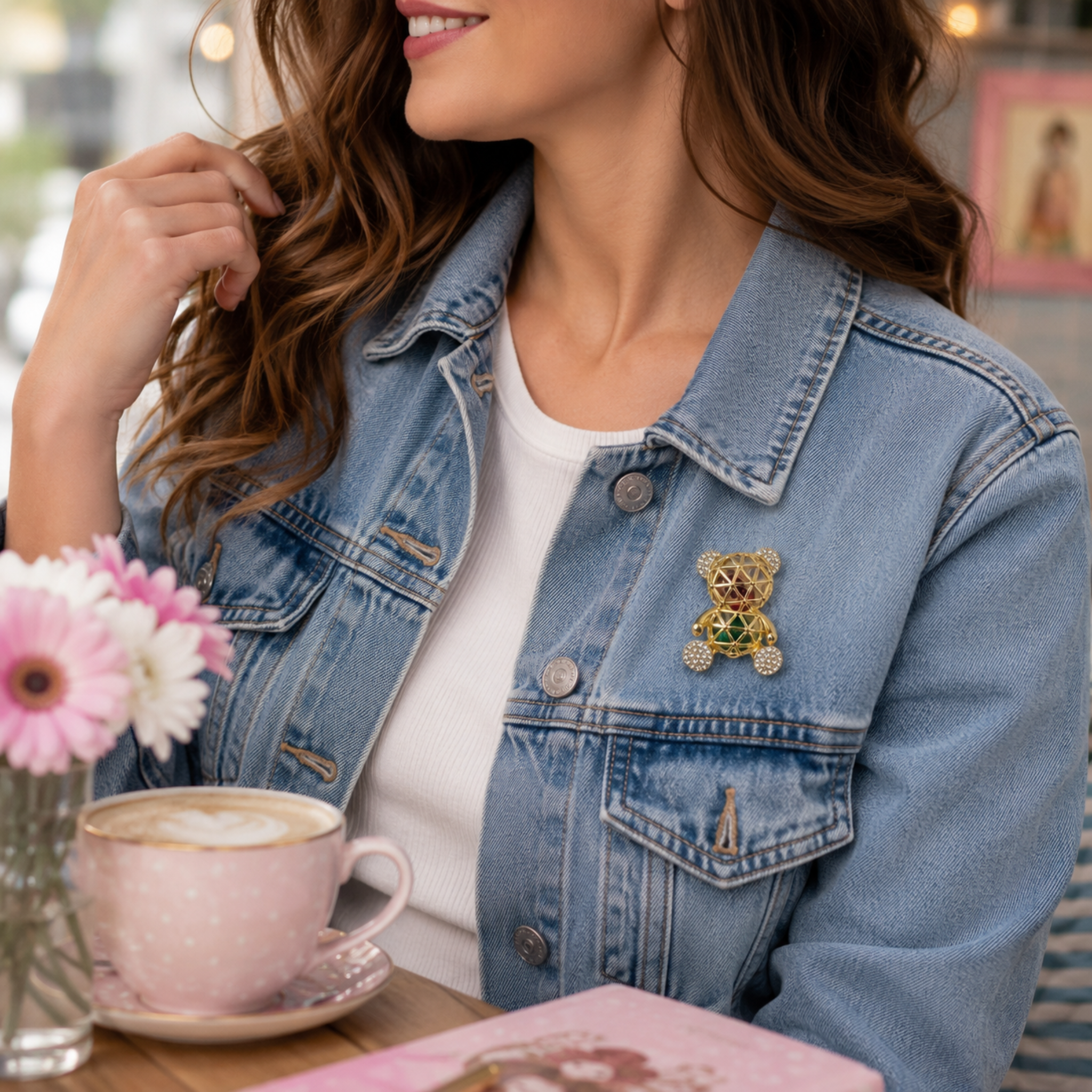 A woman wearing a light blue denim jacket with a decorative teddy bear brooch on the chest pocket, sitting at a table with a pink coffee cup and flowers.