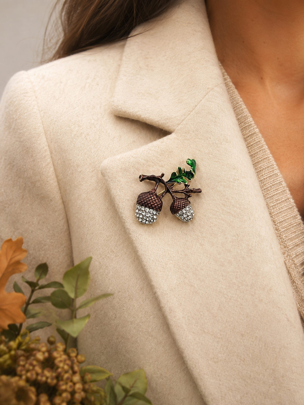 A close-up of a person wearing a cream-colored coat over a beige knit top, featuring a detailed acorn brooch with green leaves pinned to the lapel.