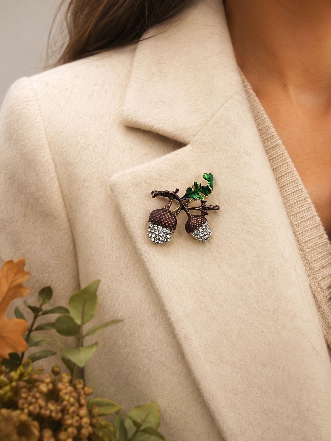 A close-up of a person wearing a cream-colored coat over a beige knit top, featuring a detailed acorn brooch with green leaves pinned to the lapel.