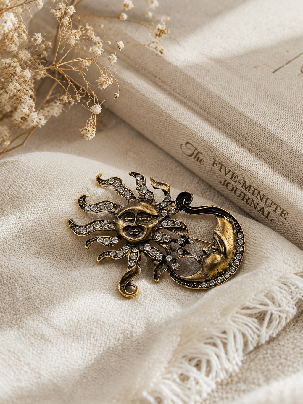 A gold-toned sun and crescent moon brooch with rhinestone accents, styled on cream fabric next to a book titled 'The FIVE-MINUTE JOURNAL' with dried flowers.