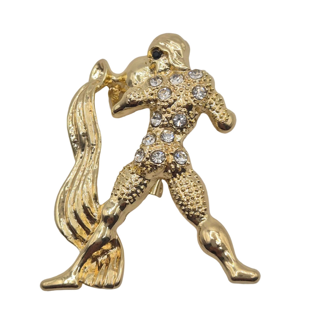 A gold-tone metal Aquarius brooch depicting a stylized figure adorned with clear rhinestones, shown on a plain white background.