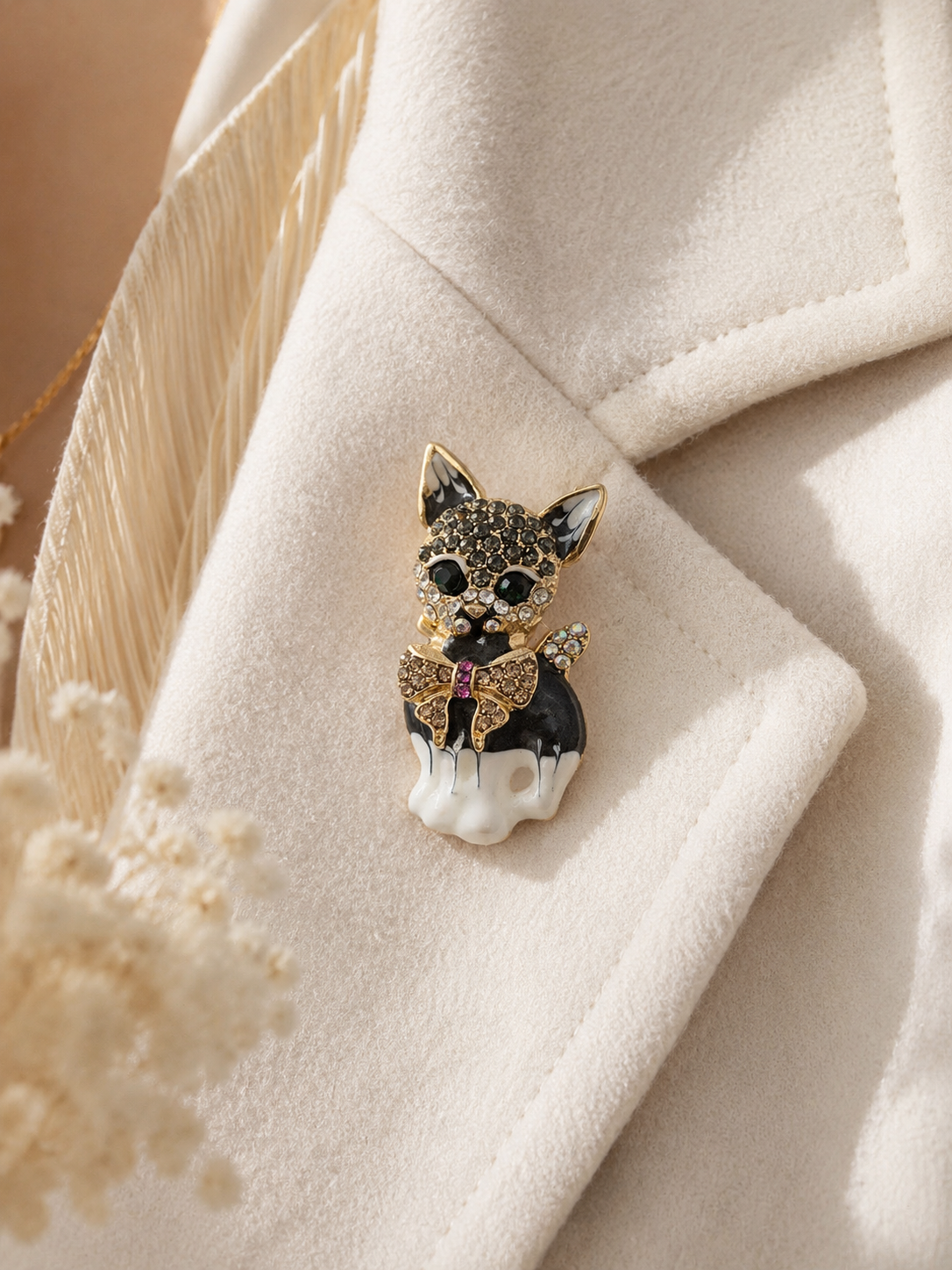 A small cat-shaped brooch with black and gold glitter details and a white paws base, pinned to a cream-colored textured coat.