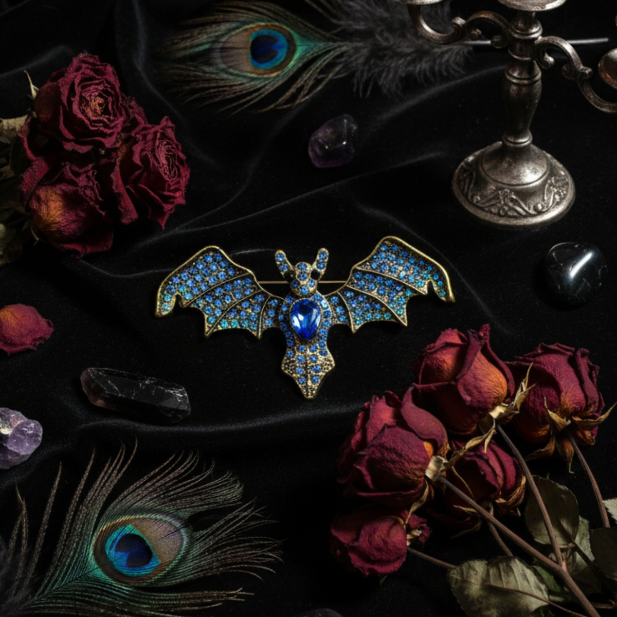 A bat-shaped brooch with blue and gold enamel detailing and a central blue gemstone, styled on black fabric with dried red roses, peacock feathers, crystals, and an antique candlestick.