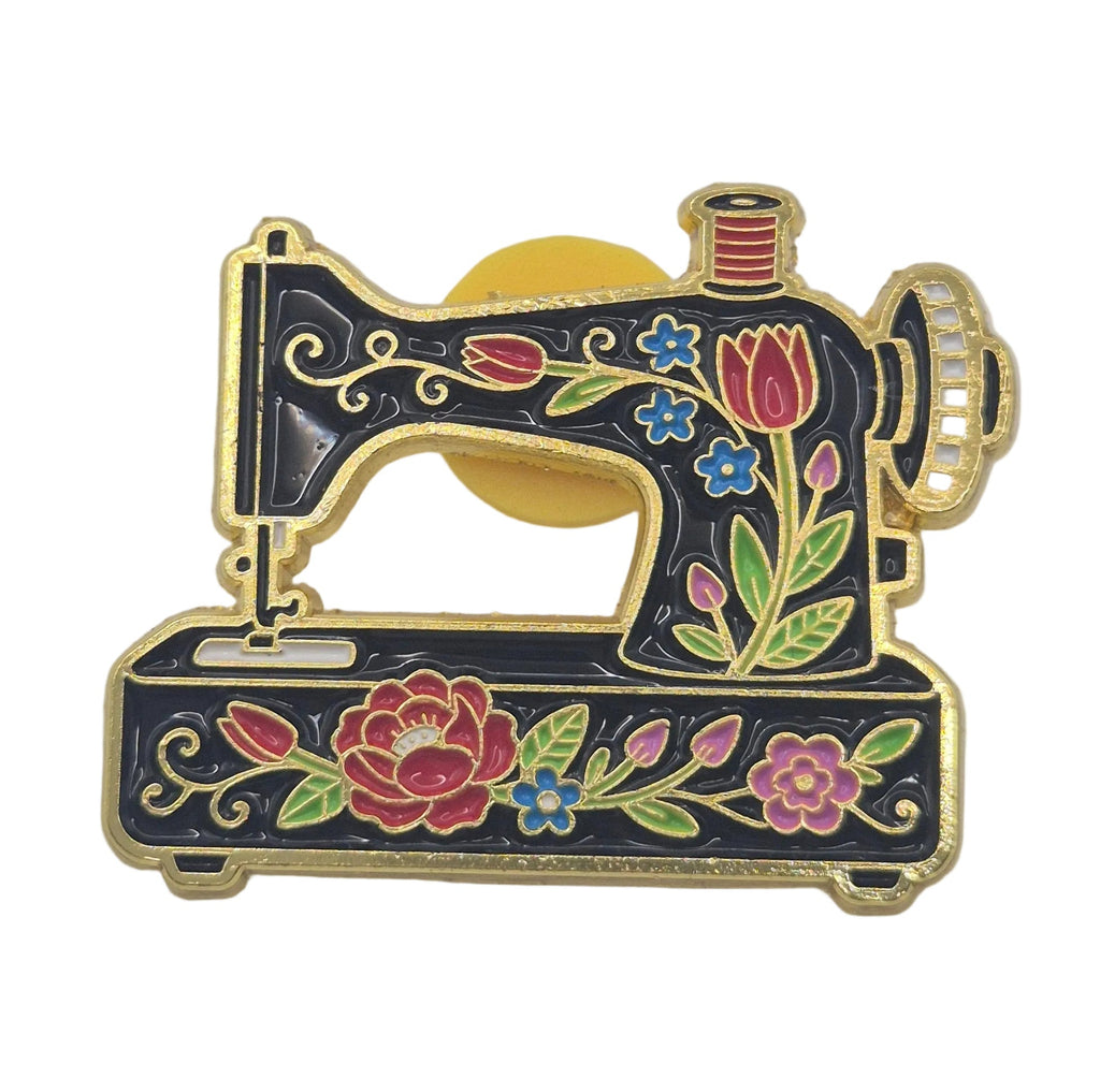 A small enamel pin shaped like a sewing machine, featuring a black body with gold-tone metal outlines and colorful floral details in pink, red, green, and blue, set against a plain white background.