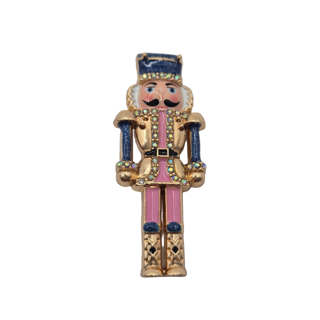 A vintage-style gold-tone nutcracker brooch with pink enamel pants, blue accents on the hat and arms, and a black moustache, shown on a plain white background.