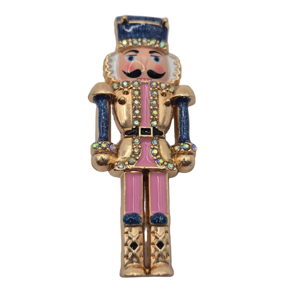 A gold-tone nutcracker figurine brooch with pink and blue enamel details, set against a plain white background.
