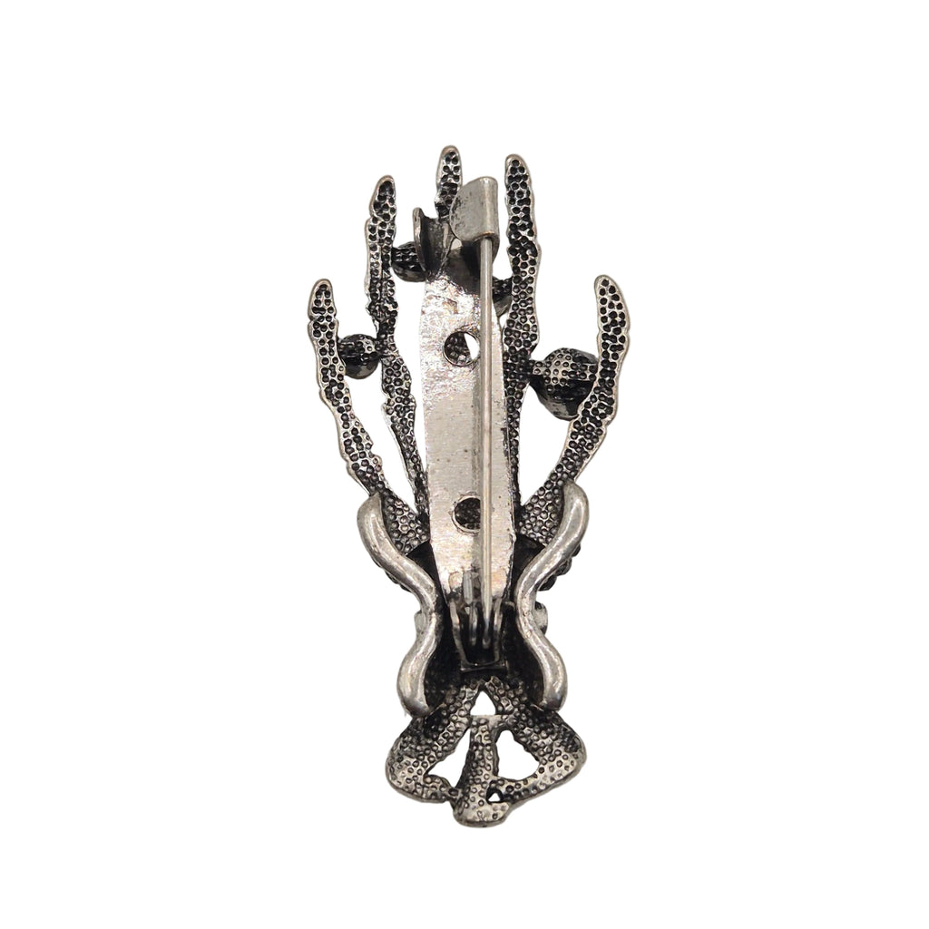 A silver-toned metal pin shaped like a skeleton hand, showing reverse side against a plain white background.