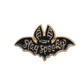 A black and gold enamel lapel pin shaped like a bat with the words 'Stay spooky' written across it, shown on a plain white background.