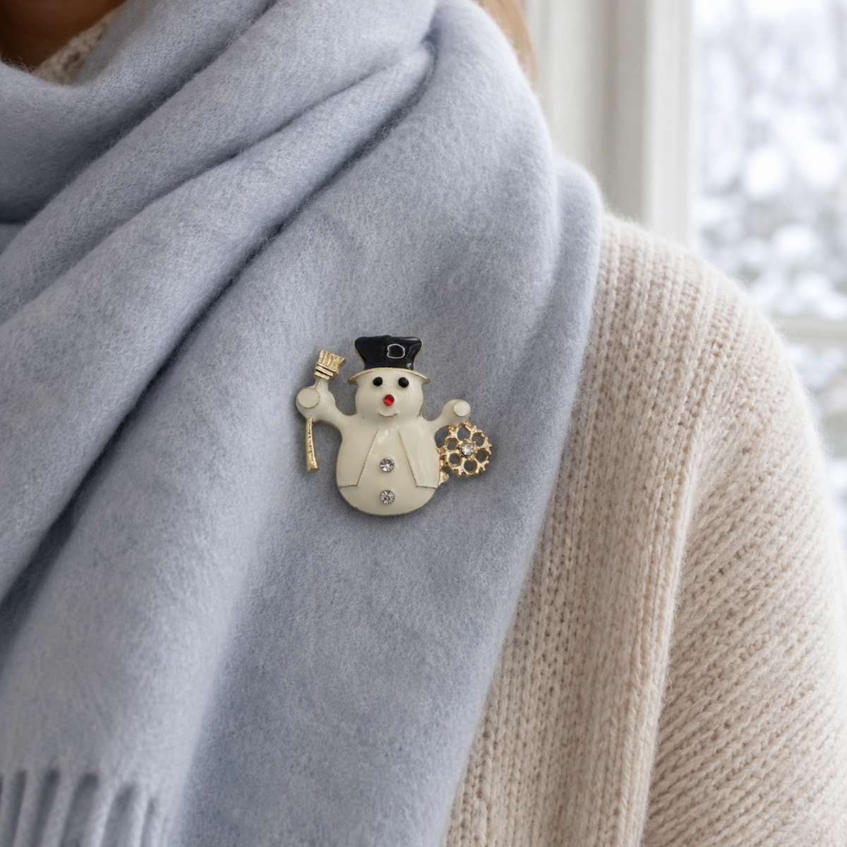 A close-up of a person wearing a light blue knit scarf and a cream knit sweater, with a small snowman brooch pinned to the scarf. The snowman has a black hat, gold accents, and a small red nose.