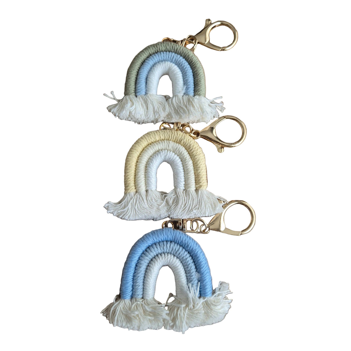 Three small woven rainbow keychains with tassels and gold-tone clasp hardware, arranged vertically on a plain white background. The keychains feature pastel green, blue, and white rainbows, pastel yellow, cream, and white rainbows, and light blue, white, and cream rainbows.