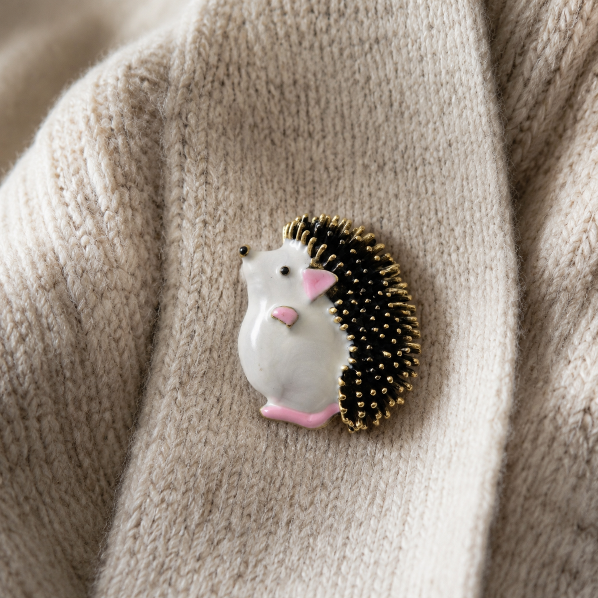 A small hedgehog-shaped brooch pinned onto a beige knit sweater. The brooch features a white body, pink ears and feet, and a black-and-gold spiny back.