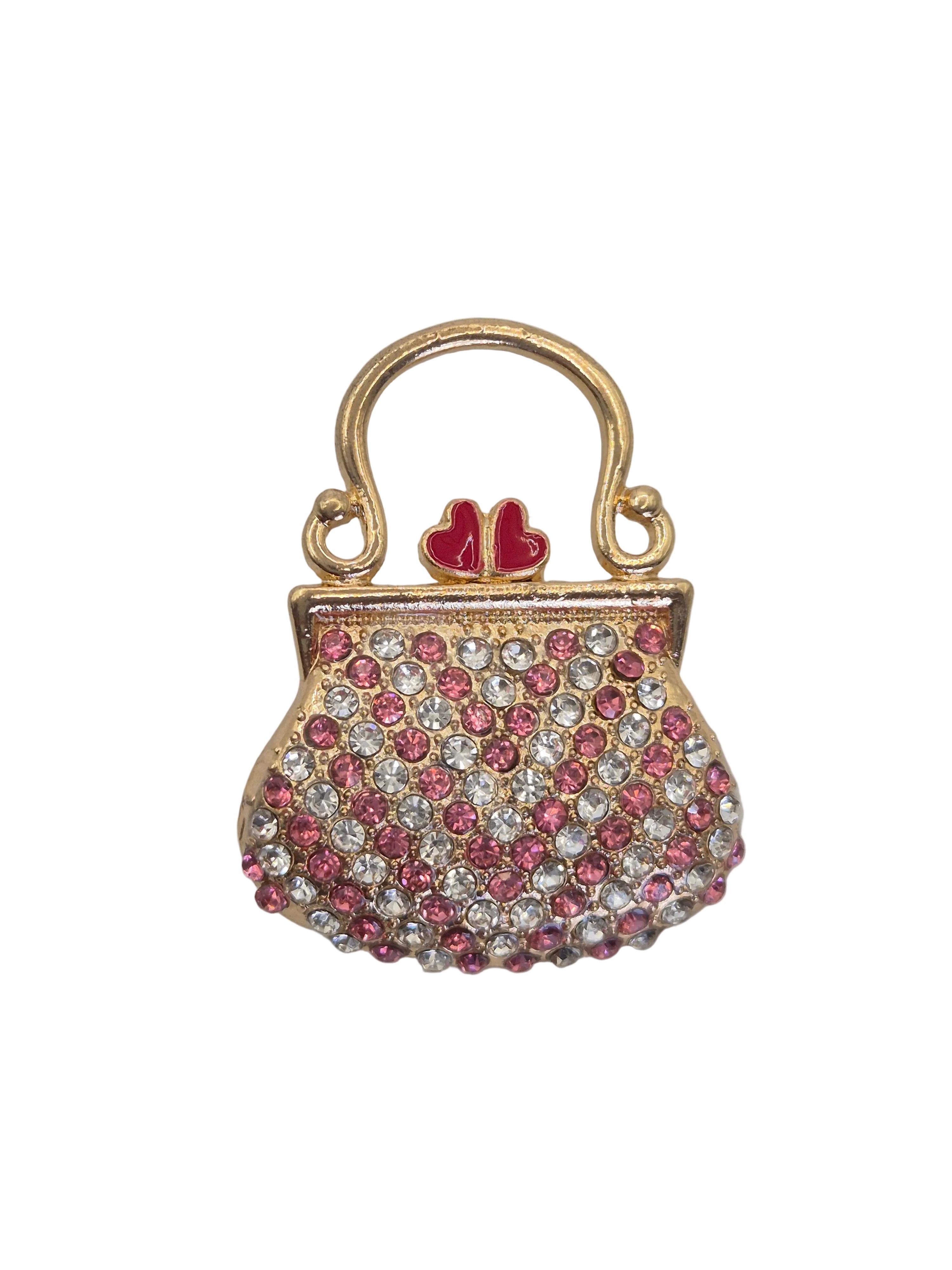 A gold-tone purse-shaped brooch embellished with pink and clear rhinestones, featuring a small red heart detail on the handle, set against a plain light gray background.