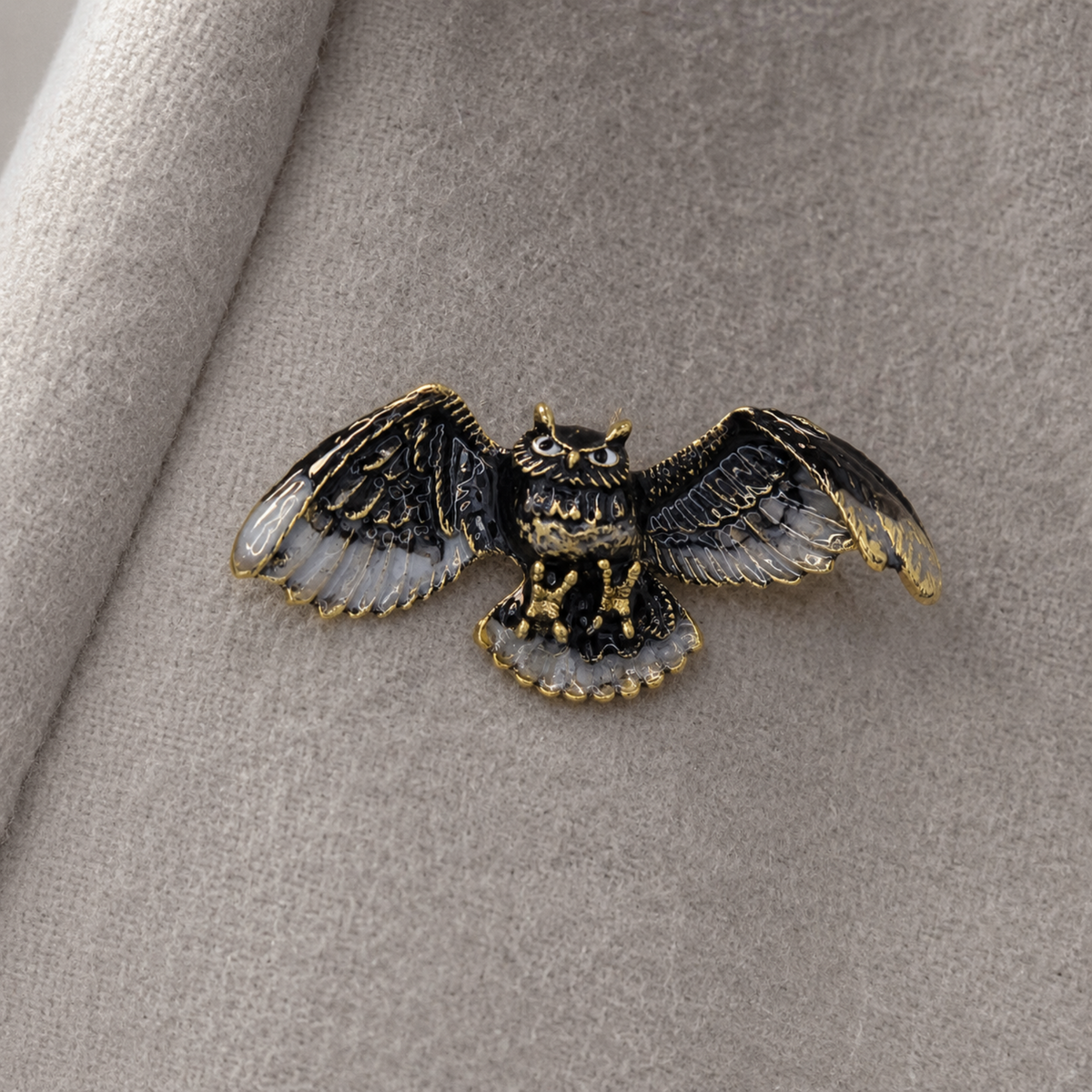 A detailed owl brooch with black and gold enamel, featuring spread wings and a textured beak, resting on a soft gray fabric background.