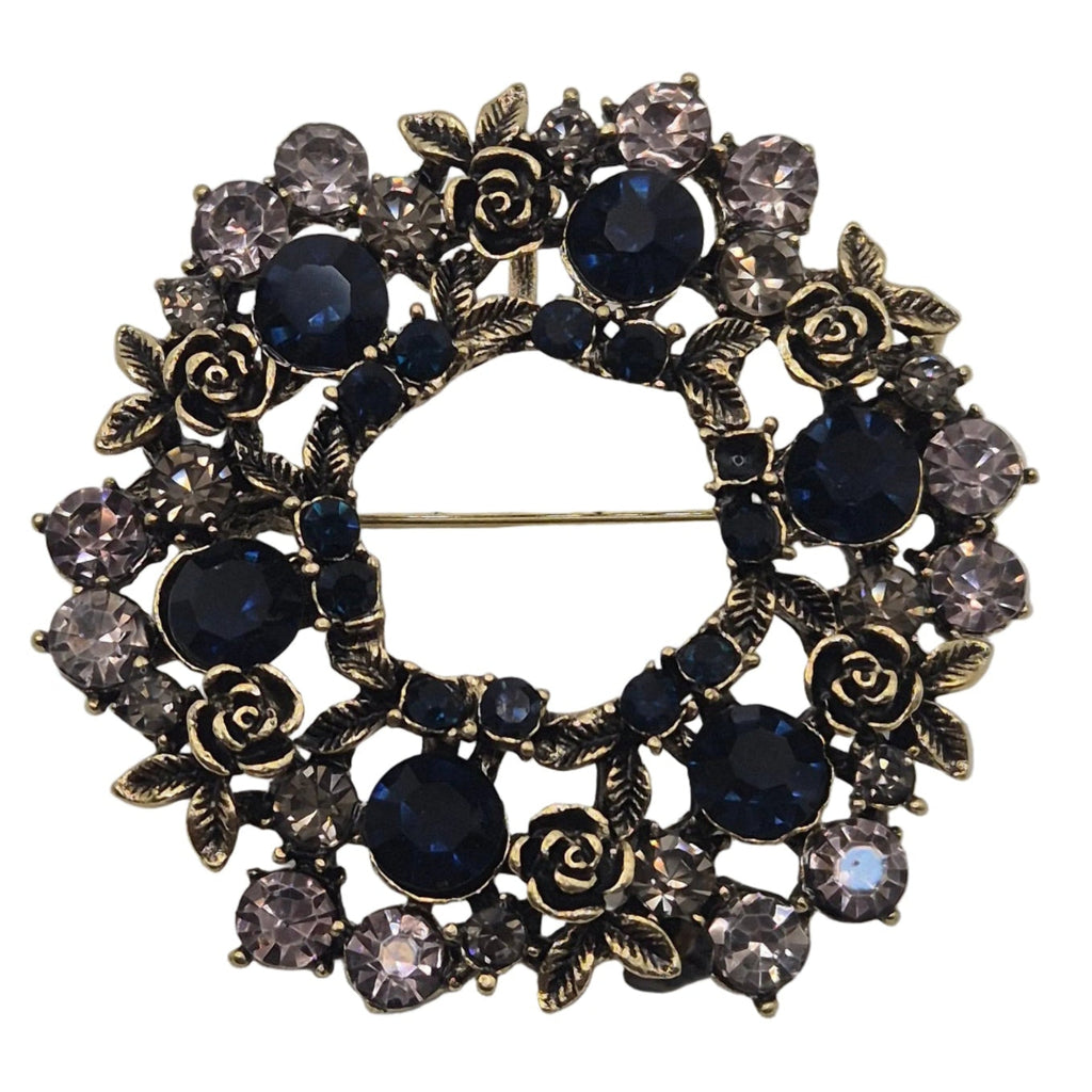 A circular brooch featuring dark blue faceted crystals, rose-shaped crystals, and antique bronze-toned metal details, set against a plain white background.