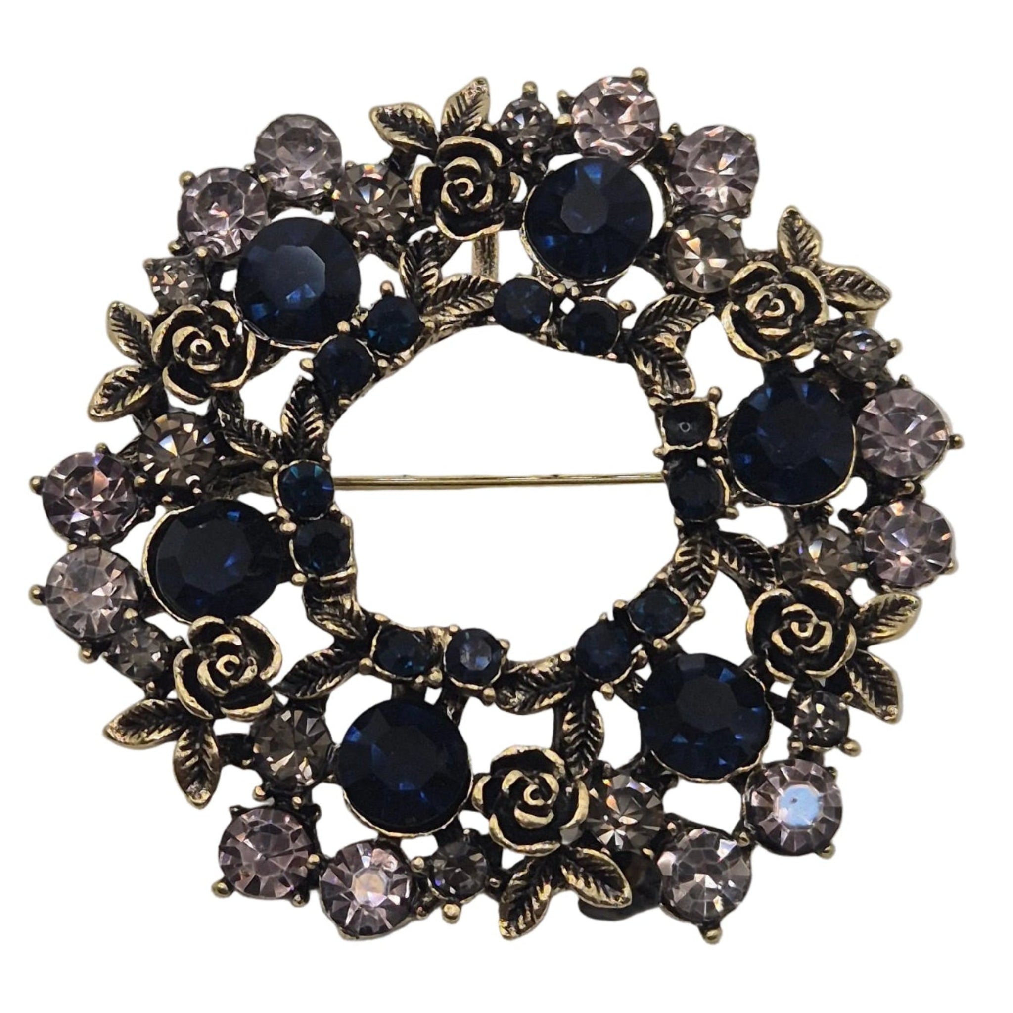 A circular brooch featuring dark blue faceted crystals, rose-shaped crystals, and antique bronze-toned metal details, set against a plain white background.