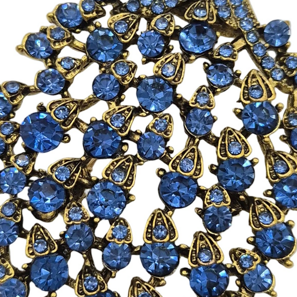 A close-up of a vintage-style peacock brooch featuring round and teardrop-cut blue rhinestones set in an antique gold-tone metal frame.