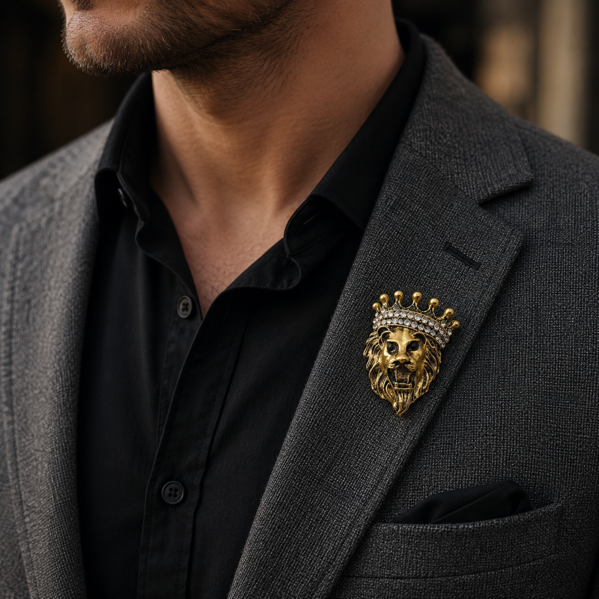 A close-up of a man wearing a textured dark gray blazer over a black button-down shirt, featuring a gold lion head and crown lapel pin on his chest.