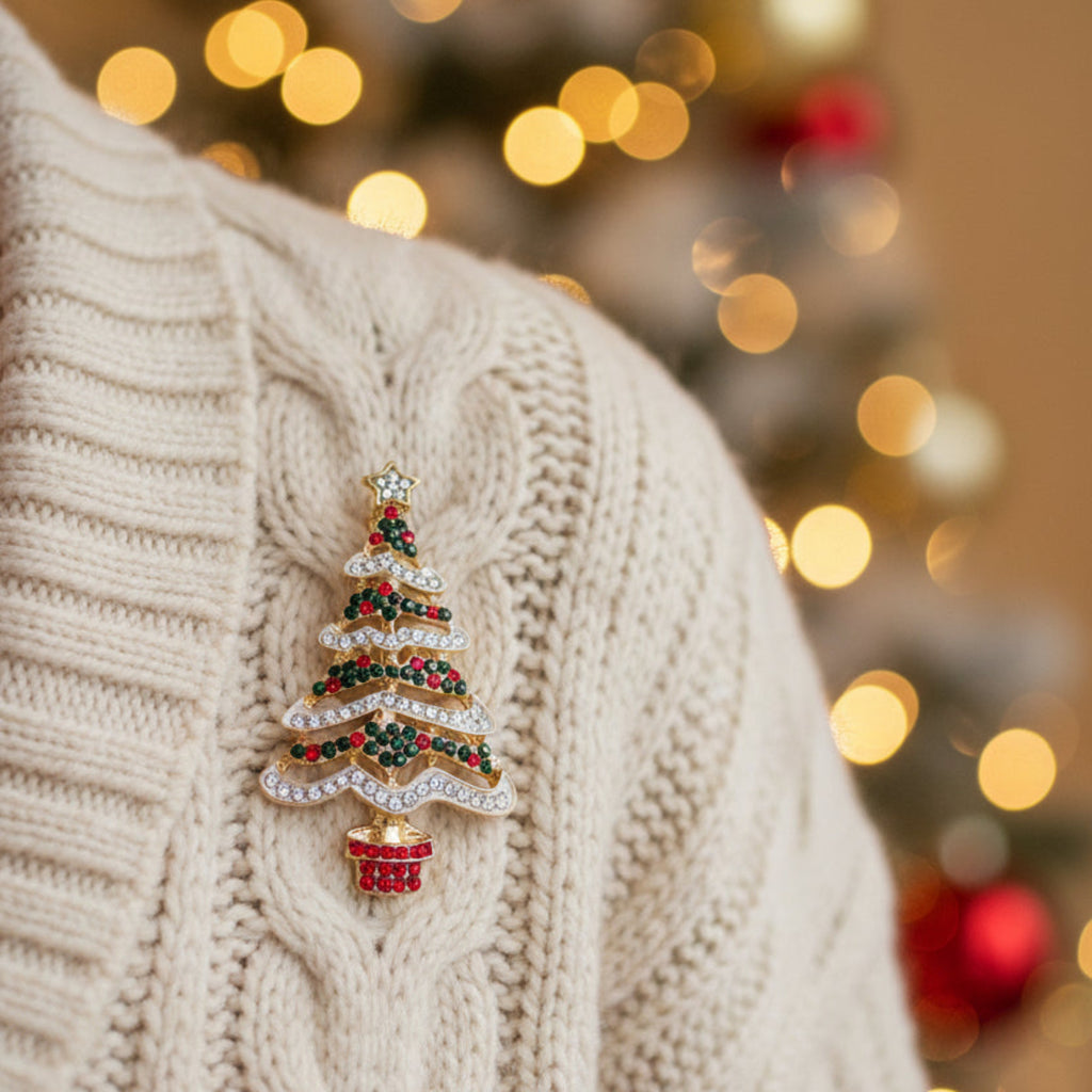 A gold Christmas tree brooch decorated with clear rhinestones, black and red rhinestones, and a small gold star on top, set against a plain light gray background.