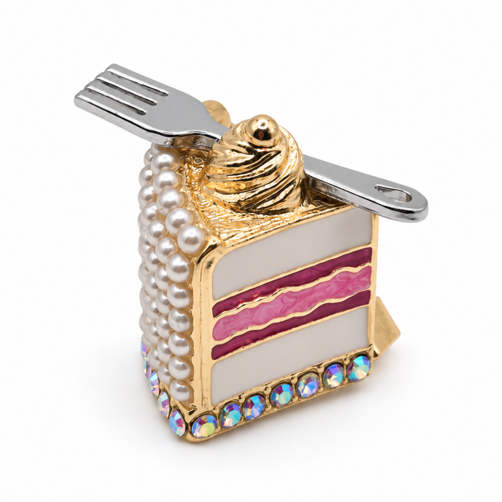 A novelty cake slice brooch with a gold-tone metal base, white faux-pearl sides, pink icing stripes, a swirl of gold frosting on top, and a silver fork resting across the upper portion. The charm is decorated with iridescent rhinestones along the bottom edge, set against a plain white background.