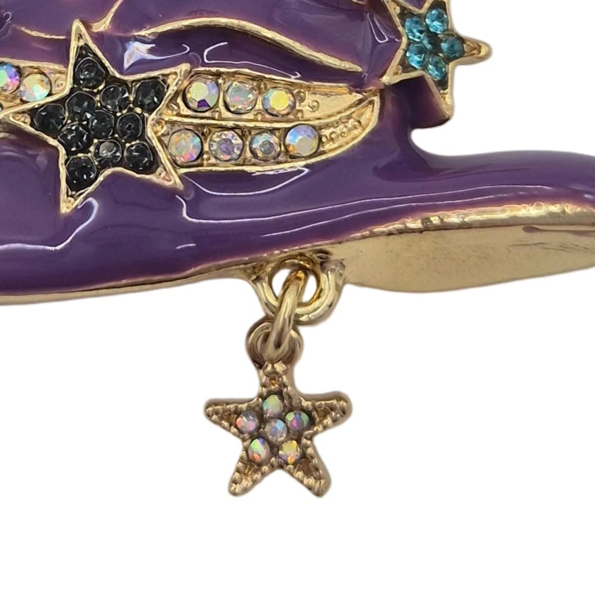 A close-up of a purple and gold enamel brooch featuring star and shooting star motifs with iridescent rhinestones, plus a dangling gold star charm on a white background.