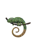 A decorative lizard or chameleon-shaped brooch embellished with green rhinestones and a curled tail paved with gold and iridescent rhinestones, shown on a plain white background.