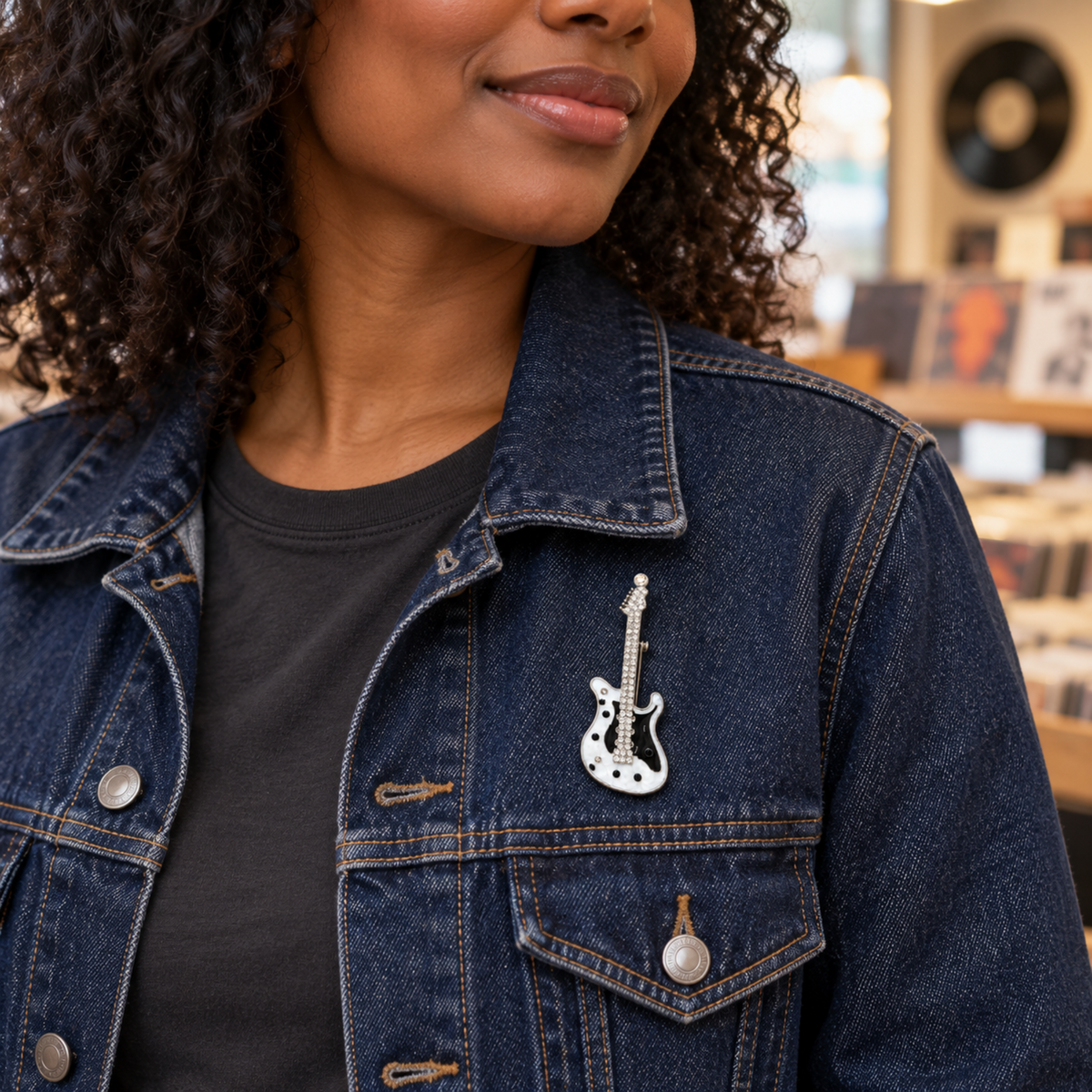 A close-up of a person wearing a dark blue denim jacket over a black t-shirt, featuring a small silver guitar-shaped lapel pin on the chest pocket.