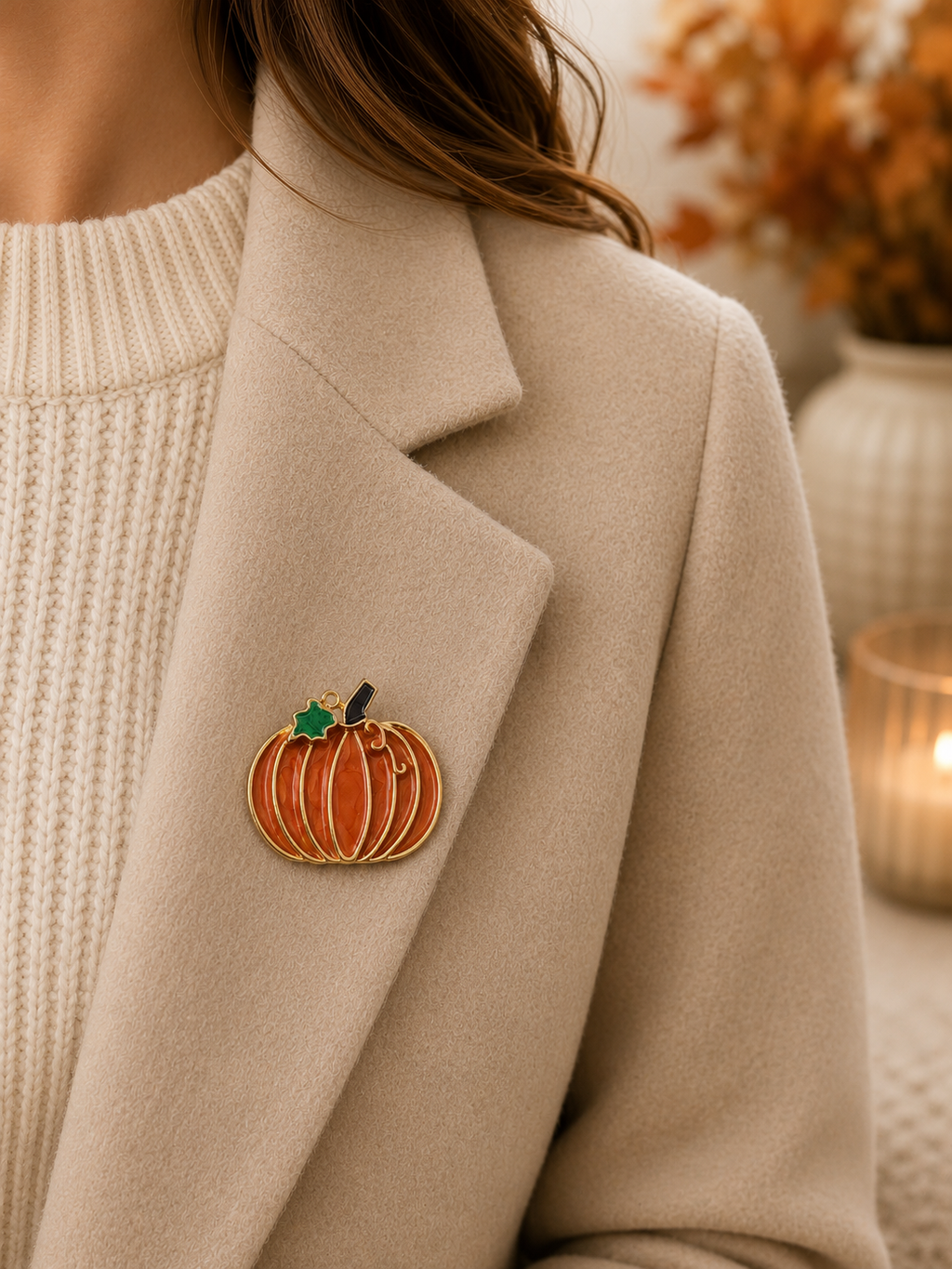 A close-up of a person wearing a beige coat over a cream knit sweater, with a small pumpkin-shaped brooch pinned to the coat lapel. The brooch is orange with gold trim, a green leaf, and a dark stem.