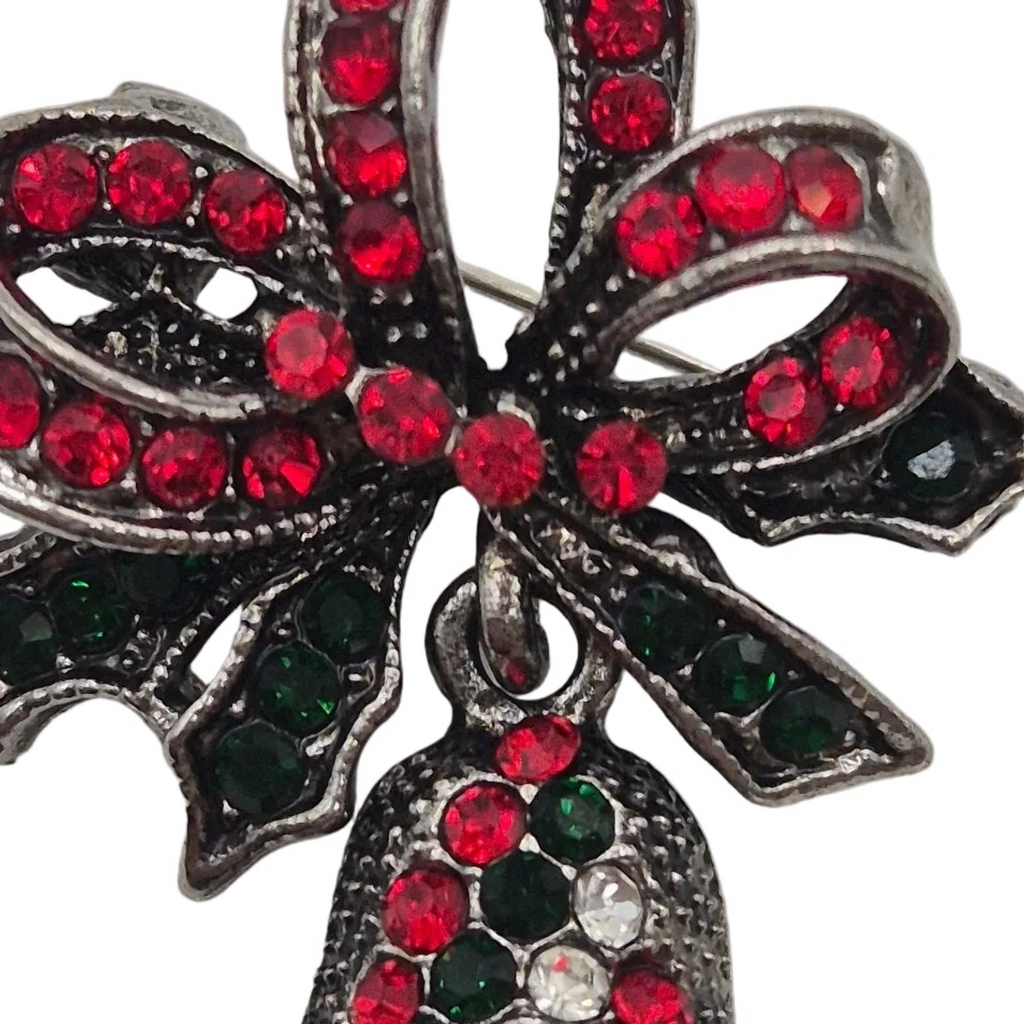 A close-up of a silver-toned Christmas ornament featuring a bow and holly design, embellished with red, dark green, and clear rhinestones on a white background.