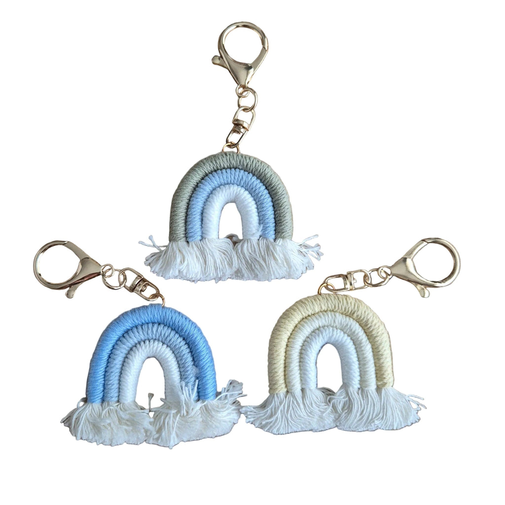 Three small rainbow keychains with tassels arranged on a plainwhitebackground. Each keychain has a gold-tone clasp and a wrapped rainbow design in soft pastel tones: one blue and white, one light blue and white, and one cream and white.