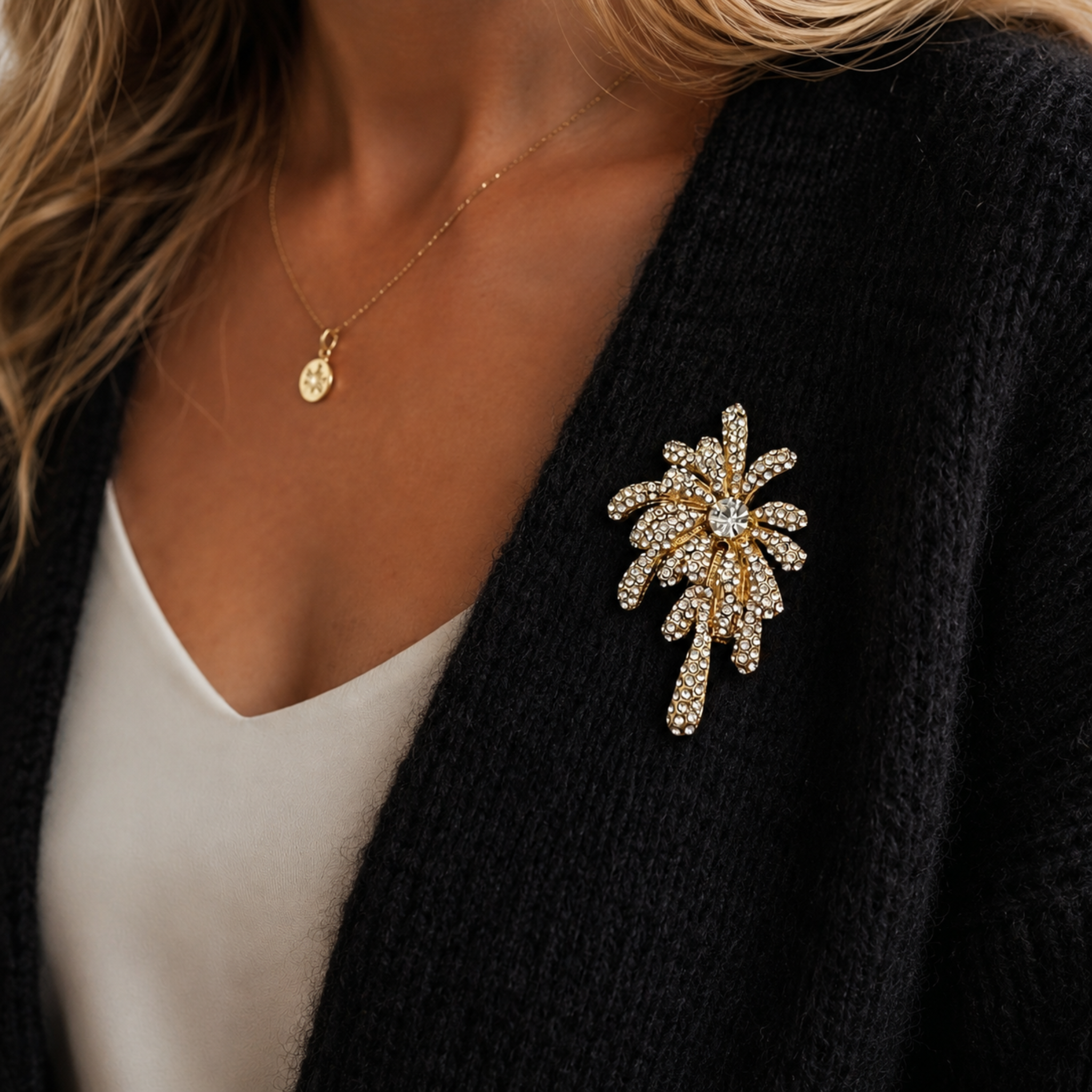A gold-toned firework brooch embellished with clear crystals, pinned to a black knit cardigan worn over a white top.